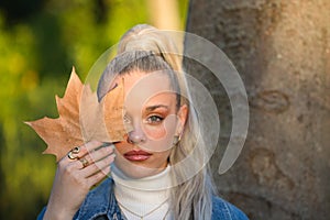 Pretty young blonde woman with a ponytail in her hair covers one eye with a dry leaf. Focus on the leaf