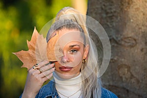 Pretty young blonde woman with a ponytail in her hair covers one eye with a dry leaf. Focus on the leaf