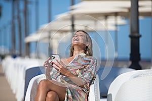 Pretty young blonde woman sitting on a terrace on the promenade. She consults her phone, social networks and takes a selfie. The
