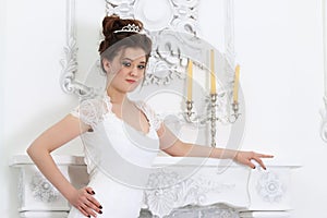 Pretty young bride in white dress poses near fireplacewith cand