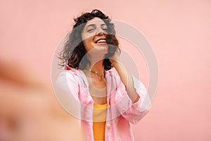 Pretty young brunette caucasian woman takes selfies looking at camera on pink background.