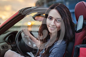 Pretty young brunette woman driving luxury red cabriolet car at the sunset.