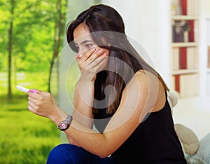 Pretty young brunette woman holding pregnancy home test, shocked facial expression covering mouth with hand, bookshelves