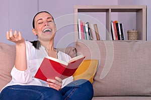 Pretty young brunette woman laughing reading a book sitting on the sofa at home.