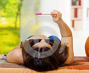 Pretty young brunette woman lying down holding up pregnancy home test in front, as seen from behind head, bookshelves