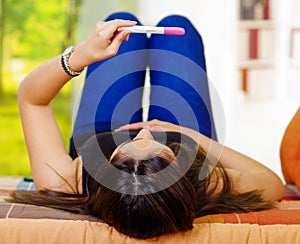 Pretty young brunette woman lying down holding up pregnancy home test in front, as seen from behind head, bookshelves