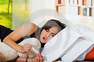 Pretty young brunette woman lying down while looking at pregnancy home test with shocked facial expression, bookshelves