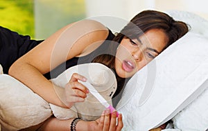 Pretty young brunette woman lying down while looking at pregnancy home test with shocked facial expression, bookshelves