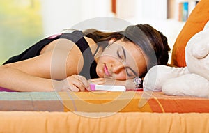 Pretty young brunette woman lying down with pregnancy home test in front, eyes closed, garden window background