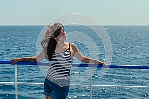 Pretty young brunette woman in a striped tank top and blue denim shorts enjoys the sea on the deck of a ship