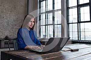 Pretty young brunette working on laptop at wooden table in spacious dark loft studio