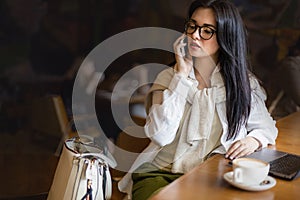 Pretty young business woman talking smartphone sitting at table in cafe with laptop and coffee cup