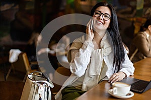 Pretty young business woman talking smartphone sitting at table in cafe with laptop and coffee cup