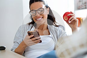 Pretty young business woman using her mobile phone while eating red apple in the office