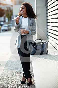 Pretty young business woman using her smart phone while drinking coffee walking down the street