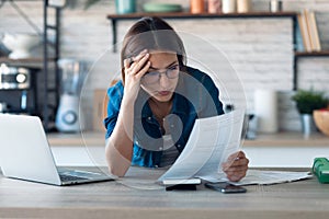 Pretty young business woman working with computer while consulting some invoices and documents in the kitchen at home