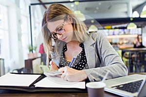 Pretty young business woman writing papers while using her mobile phone in cafe.