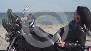 Pretty young caucasian woman in a black leather jacket and pants lying on a motorcycle near sea or river. Hobby