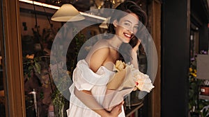 Pretty young caucasian woman with flowers smiles teeth at camera walking around city during day.