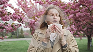 Pretty young Caucasian woman having alergy symptoms from blooming sakura tree pollen in spring. Female with pollen