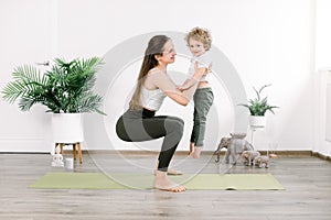 Pretty young Caucasian woman mother in sports wear does physical fitness exercises, squats on green mat, holding on