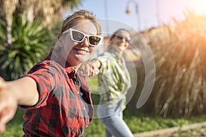 Pretty young caucasian woman in sunglasses pulling along by the hand invitingly with a happy smile as her friend on the background