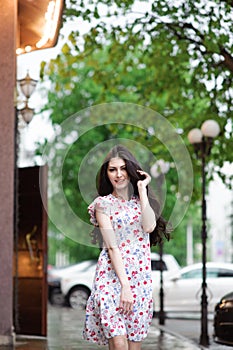 Pretty young chic caucasian girl in light dress walking along city street with green trees in spring