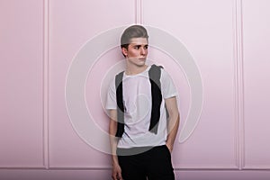 Pretty young elegant hipster man with trendy hairstyle in classic black-white clothes posing near pink wall indoors. European cute