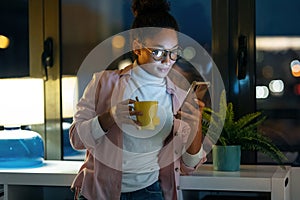 Pretty young entrepreneur woman using her smart phone while drinking a cup of coffee standing in the office