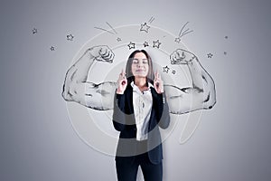 Pretty young european businesswoman with crossed fingers standing on concrete wall background with drawn muscle hands. Strength,