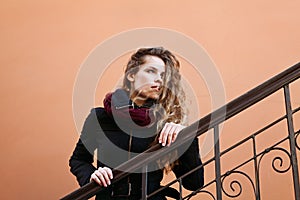 Pretty young fashion woman with long curly hair looking into the distance and posing outdoor near the wall in the street