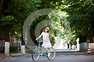 Pretty young female in white dress and straw hat riding blue bicycle middle of an empty green street