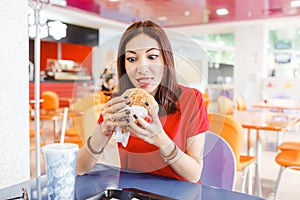 Young funny woman eating hamburger inside in fast-food restaurant