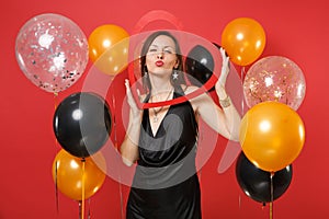 Pretty young girl in black dress holding big red wooden heart, blowing lips, sending air kiss on red background air