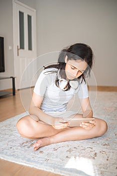 Pretty young girl on the carpet at home with a smartphone in her hand and headphones around her neck. Apartment living. Home life