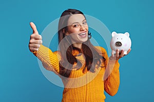 Pretty young girl in casual showing thumb up while holding white piggy bank