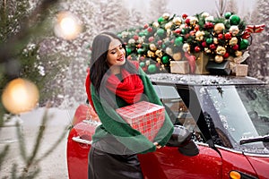 Pretty young girl is holding gift box near red car with decorated xmas tree on the roof, holiday concept.