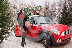 Pretty young girl is holding gift box near red car with decorated xmas tree on the roof, holiday concept.