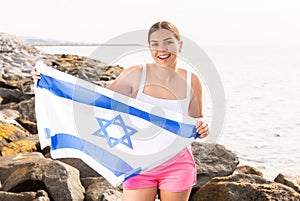 Pretty young girl holding the Israel flag in her hands on the seashore on sunny day