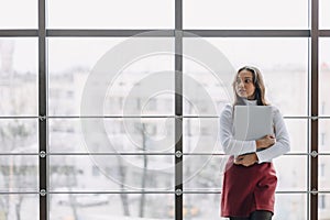 Pretty young girl with laptop by the window in office with big windows. remote work concept. freelancer working alone