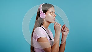 Pretty young girl listening to music, smiling, dancing in pink headphones in studio against blue background. Music