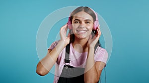 Pretty young girl with long hair listening to music, smiling, dancing hands in pink headphones in studio against blue