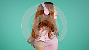 Pretty young girl with long hair listening to music, smiling, dancing with head and headphones in studio against blue