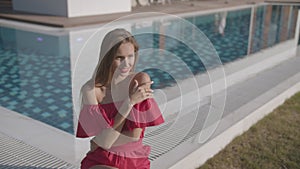 Pretty young girl in pink dress is sitting next to pool, posing and smiling