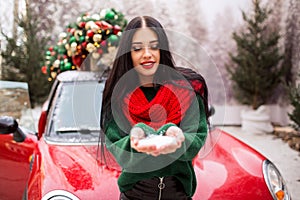 Pretty young girl is playing with snow near red car with decorated xmas tree.