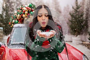 Pretty young girl is playing with snow near red car with decorated xmas tree.