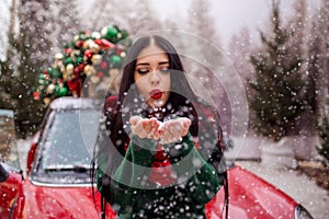 Pretty young girl is playing with snow near red car with decorated xmas tree.