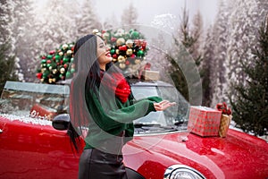 Pretty young girl is playing with snow near red car with decorated xmas tree.