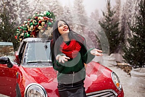 Pretty young girl is playing with snow near red car with decorated xmas tree.