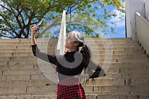 Pretty young girl in punk style taking photos with her mobile phone on a staircase in the park. Technology and beauty concept